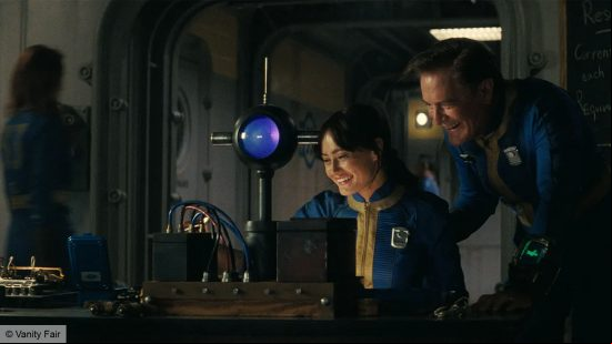 A man and a woman, dressed in blue and yellow. The man is standing, the woman is setting at a desk. There are vault control s around them. 