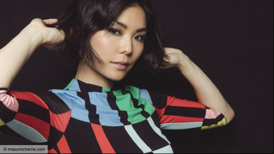 A young woman, Masumi, with short hair wearing a colorful shirt with blocks of black, blue, green, and red plays with her hair, looking into the camera